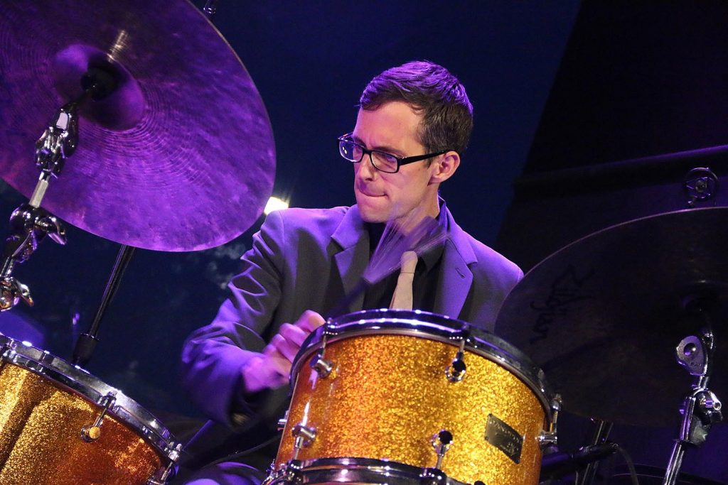 Matt Slocum performing jazz music live on the drum set wearing a suit and tie.