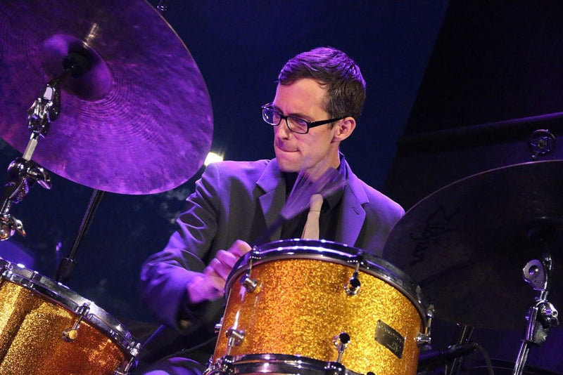 Matt Slocum performing jazz music live on the drum set wearing a suit and tie.