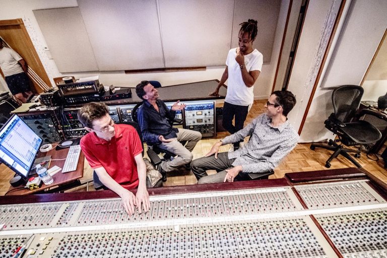 Matt Slcoum with his trio and recording person standing and talking in the recording studio with a mixing board.
